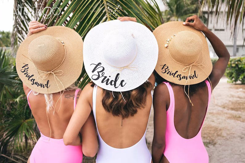 Two brown floppy hats with bridesmaid written in black and one white floppy hat with bride written in black.